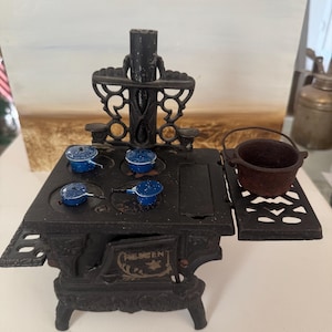 May include: A miniature black cast iron stove with the word Crescent embossed on the front door. The stove features a decorative back panel, four small blue enamel pots with lids on the burners, and a single dark brown metal pot with a handle.