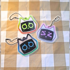 May include: Three acrylic cat-shaped keychains featuring black faces with glowing neon eyes and mouths. One keychain has bright green features, one has blue features, and one has pink features. Each includes a silver metal ball chain attachment.