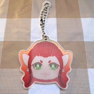 May include: A clear acrylic keychain featuring a cartoon character with red hair, green eyes, and a smiling expression. The keychain has a silver chain and a metal ring for attachment. The character's face is outlined in orange.