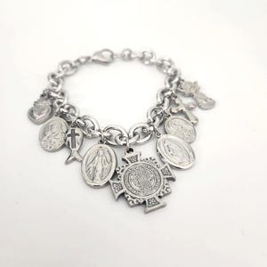 May include: A silver-toned charm bracelet featuring multiple religious pendants, including a Saint Benedict cross, several oval medals depicting figures, a heart, a fish-shaped cross, and a small angel figure attached to a thick chain link band.