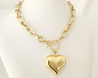 May include: A gold chain necklace with a large, puffed heart pendant. The chain is a thick, chunky style.