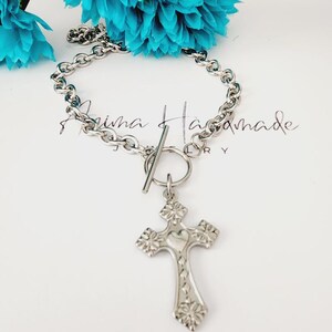 May include: A silver chain necklace with a toggle clasp and a silver cross pendant. The cross has a heart design in the center and floral details around the edges.