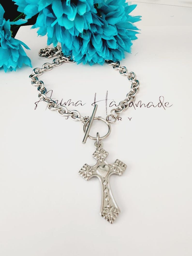 May include: A silver chain necklace with a toggle clasp and a silver cross pendant. The cross has a heart design in the center and floral details around the edges.