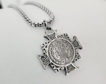 May include: A silver chain necklace with a silver pendant featuring a detailed cross with a round medallion in the center. The medallion has an image of a saint and the text "SANCTA MARIA" around the edge.