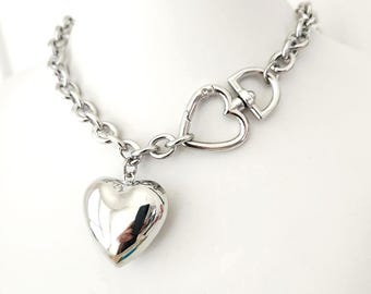May include: A silver-colored necklace with a heart-shaped pendant. The necklace features a chain design and a heart-shaped clasp. The pendant is a large, polished heart. The necklace is displayed on a white surface.