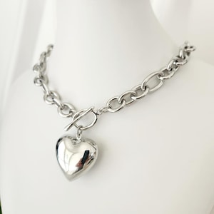 May include: A silver chain necklace with a large, heart-shaped pendant. The chain is thick and chunky, and the pendant is smooth and shiny.