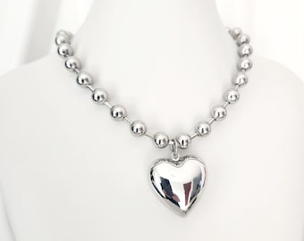 May include: A silver-colored necklace with a large, heart-shaped pendant. The necklace features a chain of small, round beads. The heart pendant is smooth and reflective, catching the light. The necklace is displayed on a white mannequin.