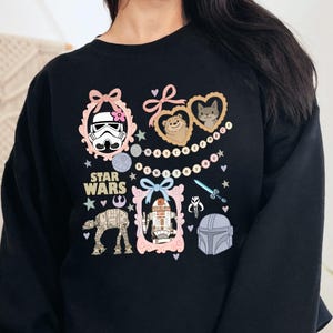 May include: Black sweatshirt featuring Star Wars characters in decorative frames. The design includes images of a Stormtrooper, Ewoks, R2-D2, and a Mandalorian helmet, with the words "Star Wars" in the center.