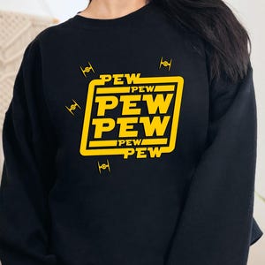 May include: Black sweatshirt with a yellow graphic that says "PEW PEW PEW" in a square. The design includes small yellow starfighter illustrations. The sweatshirt is a crew neck style.
