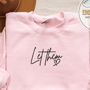 May include: Pink sweatshirt with the text "Let them" in black script with a small heart.