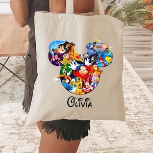 May include: A beige tote bag featuring a colorful Disney character design in the shape of Mickey Mouse's head. The bag has long handles and the name "Olivia" printed in script. The bag is held by a person.
