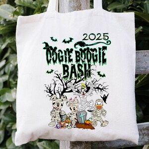 May include: Off-white tote bag with a Halloween-themed graphic. The design features the text "DOGIE BOOGIE BASH 2025" in green, along with cartoon characters dressed as mummies. A spooky tree and bats complete the design.