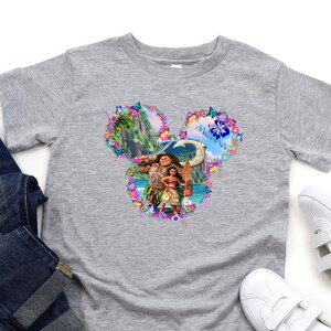Puede incluir: Una camiseta gris con un gr&aacute;fico colorido de Moana y Maui en forma de cabeza de Mickey Mouse, rodeada de elementos florales. La camiseta se combina con vaqueros azules y zapatillas blancas con rayas negras.