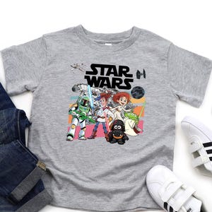 Puede incluir: Una camiseta gris de Star Wars con un dise&ntilde;o de dibujos animados colorido con personajes de la pel&iacute;cula. La imagen incluye un par de vaqueros azules y zapatillas blancas con rayas negras.