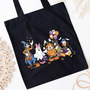 May include: Black tote bag with a Halloween design featuring Disney characters in costumes. Goofy, Minnie Mouse, Mickey Mouse, and Donald Duck are shown with pumpkins, ghosts, and balloons. The bag has two black handles.