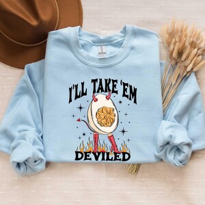 May include: A light blue sweatshirt with the text "I'll Take 'Em Deviled" in black. A cartoon illustration of a deviled egg with red devil horns and red boots is in the center of the text.