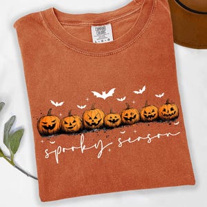 May include: Orange Comfort Colors t-shirt featuring a row of jack-o'-lanterns and white bats. The words "spooky season" are written in a cursive font below the pumpkins. A brown hat is visible in the background.