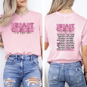 May include: A light pink t-shirt with the words "BREAST CANCER" in a pink, textured font. The front of the shirt has the text "The Fight Tour". The back of the shirt has a list of words related to breast cancer awareness.