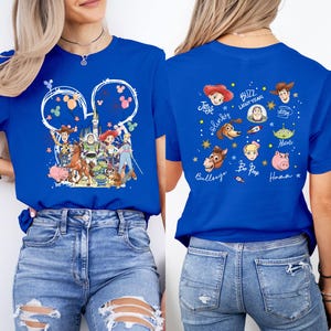 Puede incluir: Camiseta azul real con un dise&ntilde;o tem&aacute;tico de Disney. La parte delantera muestra un collage de personajes de Toy Story dentro de un contorno de oreja de Mickey Mouse. La parte trasera muestra ilustraciones y nombres de personajes individuales.