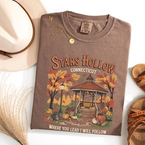 May include: A brown t-shirt with the words "Stars Hollow Connecticut" and a gazebo graphic. The shirt is paired with a beige hat, gold necklace, and brown sandals. The graphic includes pumpkins and fall foliage.