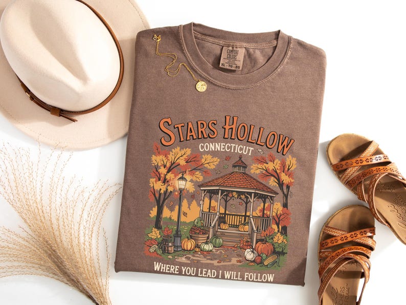 May include: A brown t-shirt with the words "Stars Hollow Connecticut" and a gazebo graphic. The shirt is paired with a beige hat, gold necklace, and brown sandals. The graphic includes pumpkins and fall foliage.