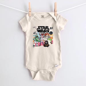 Puede incluir: Body de beb&eacute; color crema con un gr&aacute;fico colorido de Star Wars. El dise&ntilde;o incluye el logotipo de Star Wars e ilustraciones de dibujos animados de personajes como Darth Vader y un Stormtrooper. El body est&aacute; colgado de un tendedero.