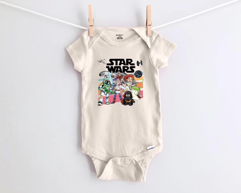 Puede incluir: Body de beb&eacute; color crema con un gr&aacute;fico colorido de Star Wars. El dise&ntilde;o incluye el logotipo de Star Wars e ilustraciones de dibujos animados de personajes como Darth Vader y un Stormtrooper. El body est&aacute; colgado de un tendedero.