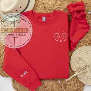 May include: A red sweatshirt with a white embroidered face design and the text "His Sally" on the sleeve. The sweatshirt is displayed on a woven surface with a straw hat and fan.
