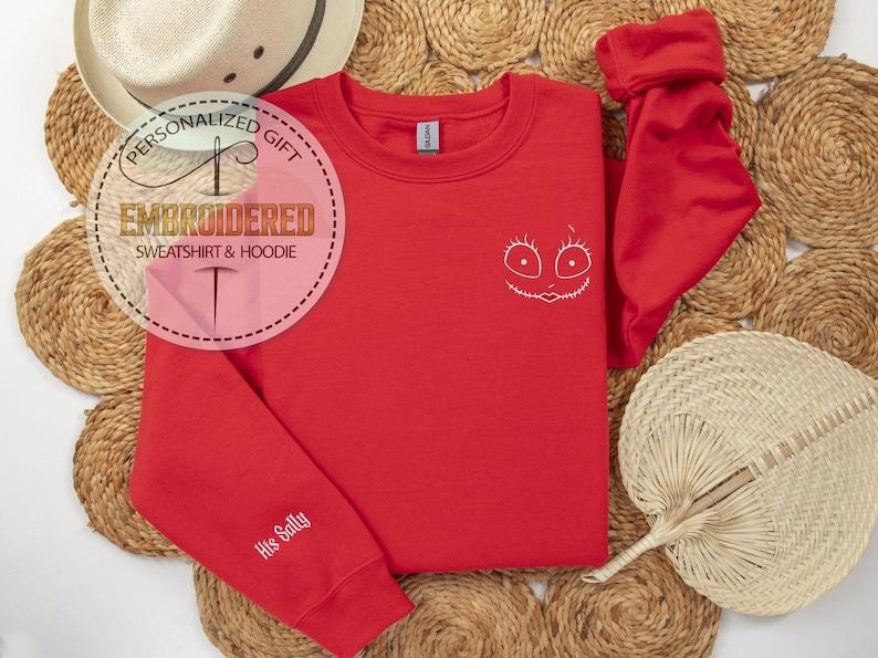May include: A red sweatshirt with a white embroidered face design and the text "His Sally" on the sleeve. The sweatshirt is displayed on a woven surface with a straw hat and fan.