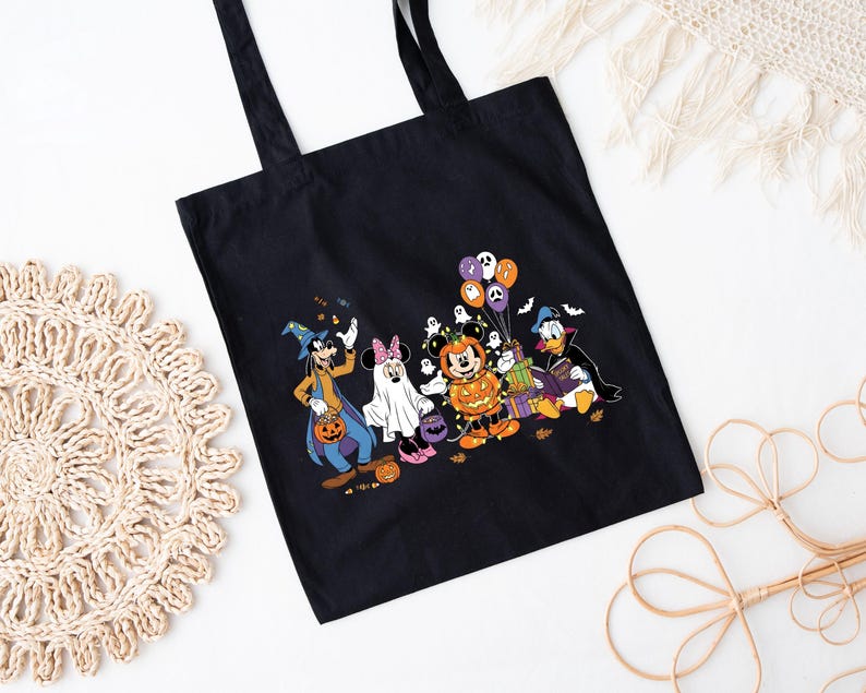 May include: Black tote bag with a Halloween design featuring Disney characters in costumes. Goofy, Minnie Mouse, Mickey Mouse, and Donald Duck are shown with pumpkins, ghosts, and balloons. The bag has two black handles.