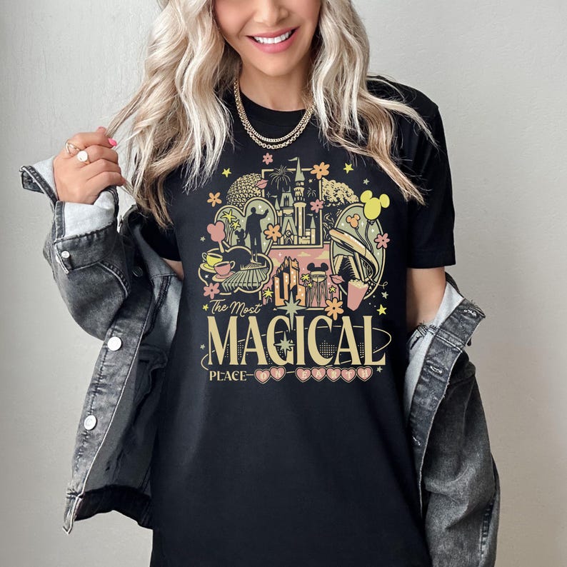 Puede incluir: Camiseta negra con un gr&aacute;fico colorido que presenta un castillo, orejas de Mickey Mouse y otras im&aacute;genes de parques tem&aacute;ticos. El texto de la camiseta dice "The Most MAGICAL PLACE". El dise&ntilde;o incluye elementos amarillos, rosas y verdes.
