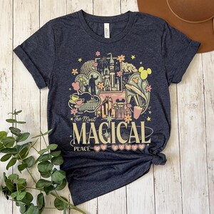 Puede incluir: Una camiseta azul marino con un gr&aacute;fico colorido que dice "The Most Magical Place". El dise&ntilde;o incluye un castillo, orejas de Mickey Mouse y otros elementos de parques tem&aacute;ticos. La camiseta est&aacute; anudada en la parte inferior.