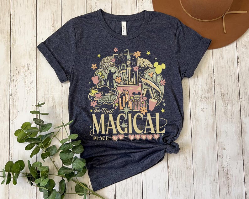 Puede incluir: Una camiseta azul marino con un gr&aacute;fico colorido que dice "The Most Magical Place". El dise&ntilde;o incluye un castillo, orejas de Mickey Mouse y otros elementos de parques tem&aacute;ticos. La camiseta est&aacute; anudada en la parte inferior.