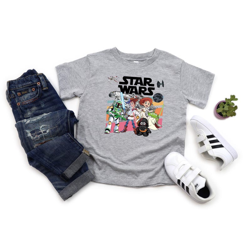 Puede incluir: Una camiseta gris de Star Wars con un dise&ntilde;o de dibujos animados colorido con personajes de la pel&iacute;cula. La imagen incluye un par de vaqueros azules y zapatillas blancas con rayas negras.