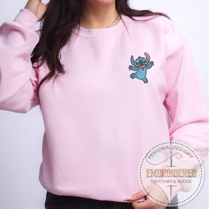 May include: A light pink sweatshirt with a small embroidered image of a blue cartoon character. The character is smiling and has large ears. The sweatshirt has a round neckline and long sleeves. The text "PERSONALIZED GIFT" and "EMBROIDERED SWEATSHIRT & HOODIE" is printed on a circular sticker.