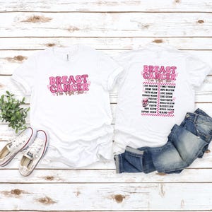 May include: Two white t-shirts with the words "BREAST CANCER" in pink, with a pink ribbon design. One shirt has the text on the front, the other on the back with a list of related words. A pair of white sneakers and blue jeans are also in the image.