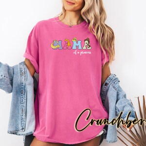 K&ouml;nnte beinhalten: Pinkes T-Shirt mit einem Aufdruck von Disney-Prinzessinnen, die "Mama of a Princess" buchstabieren.