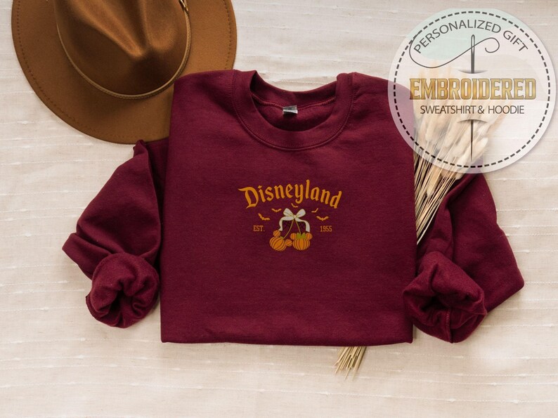 Pode incluir: Camisola bord&ocirc; com gola redonda com a palavra "Disneyland" bordada em dourado, junto com ab&oacute;boras e morcegos. A camisola tem mangas compridas e decote redondo. Um chap&eacute;u castanho est&aacute; no fundo.