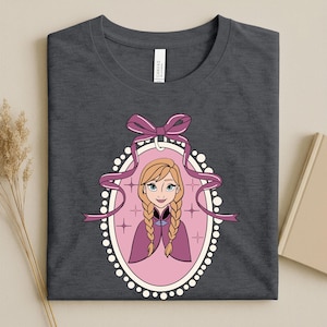 May include: Dark gray t-shirt featuring a cartoon illustration of a character with blonde braids, wearing a purple cape, inside a pink oval frame with a white border. A purple bow and ribbons are at the top of the frame.