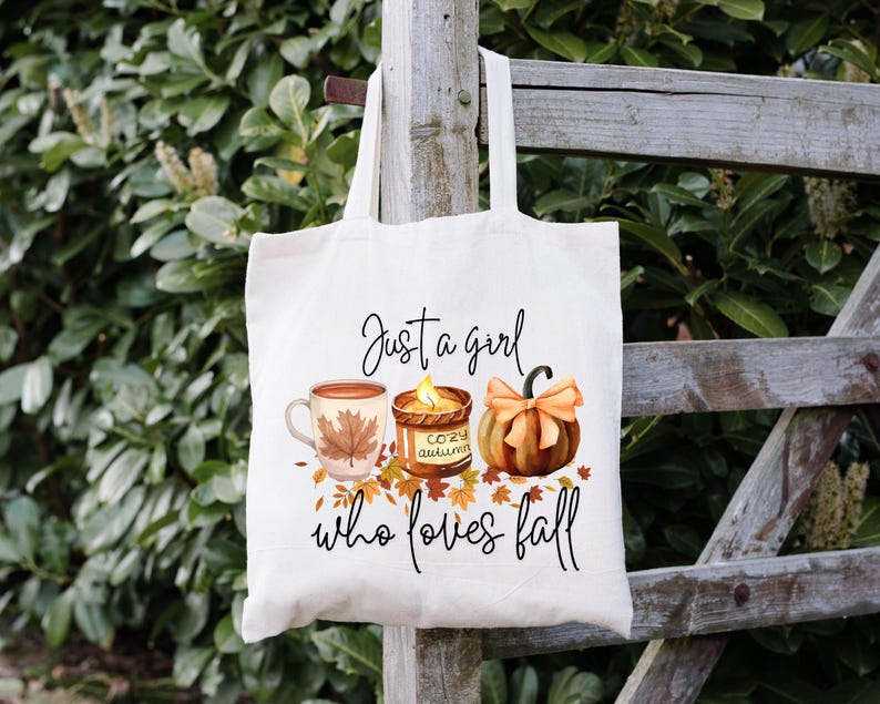 May include: Cream-colored tote bag with the text "Just a girl who loves fall." The bag features illustrations of a mug with a leaf, a candle, and a pumpkin. The design is in warm autumn colors.