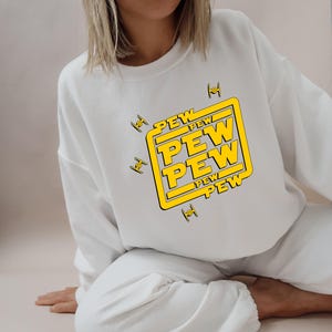 May include: White sweatshirt with a yellow and black graphic that says "PEW PEW PEW" inside a yellow square. The design includes small black and yellow starfighter illustrations. The sweatshirt is a casual, comfortable style.