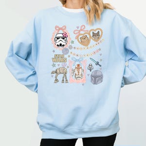 May include: Light blue sweatshirt featuring Star Wars characters and icons. The design includes illustrations of a Stormtrooper, AT-AT walker, R2-D2, and Mandalorian helmet, framed with pink bows and hearts. The words "Star Wars" are also visible.