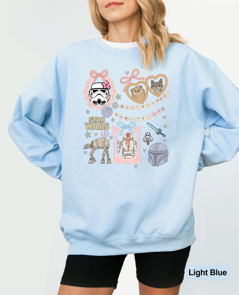 May include: Light blue sweatshirt featuring Star Wars characters and icons. The design includes illustrations of a Stormtrooper, AT-AT walker, R2-D2, and Mandalorian helmet, framed with pink bows and hearts. The words "Star Wars" are also visible.