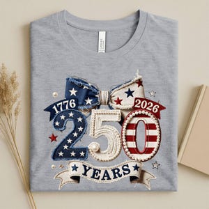 May include: A heather gray t-shirt featuring a graphic of a large bow with a patriotic theme. The design includes the numbers 1776 and 2026, the text 250 YEARS, and stars and stripes patterns in red, white, and blue with pearl-like accents.
