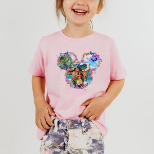 Puede incluir: Una camiseta rosa claro con un gr&aacute;fico colorido de personajes y paisajes de una popular pel&iacute;cula de animaci&oacute;n. El dise&ntilde;o tiene forma de orejas de Mickey Mouse y est&aacute; rodeado de detalles florales. La camiseta la lleva un ni&ntilde;o peque&ntilde;o.