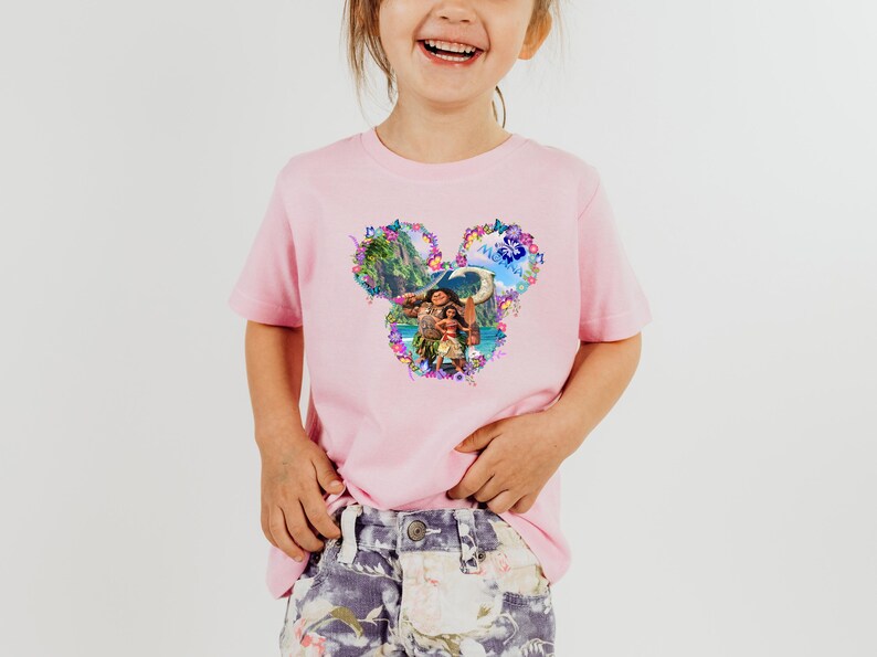 Puede incluir: Una camiseta rosa claro con un gr&aacute;fico colorido de personajes y paisajes de una popular pel&iacute;cula de animaci&oacute;n. El dise&ntilde;o tiene forma de orejas de Mickey Mouse y est&aacute; rodeado de detalles florales. La camiseta la lleva un ni&ntilde;o peque&ntilde;o.