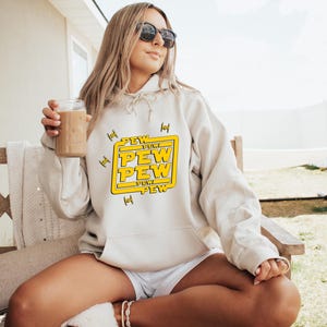 May include: Cream-colored hoodie with the text "PEW PEW" in a yellow box, with starfighter graphics. The hoodie is worn by a person wearing sunglasses, sitting on a wooden bench, holding a glass of a brown beverage.
