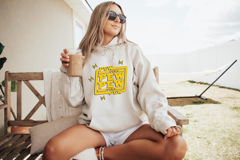 May include: Cream-colored hoodie with the text "PEW PEW" in a yellow box, with starfighter graphics. The hoodie is worn by a person wearing sunglasses, sitting on a wooden bench, holding a glass of a brown beverage.