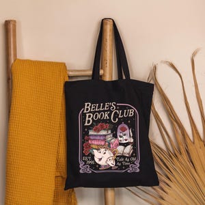 May include: Black tote bag with the text "Belle's Book Club" and a graphic of books, a teapot, and a rose. The bag is hanging on a wooden ladder with a yellow blanket. Dried palm leaves are on the right.