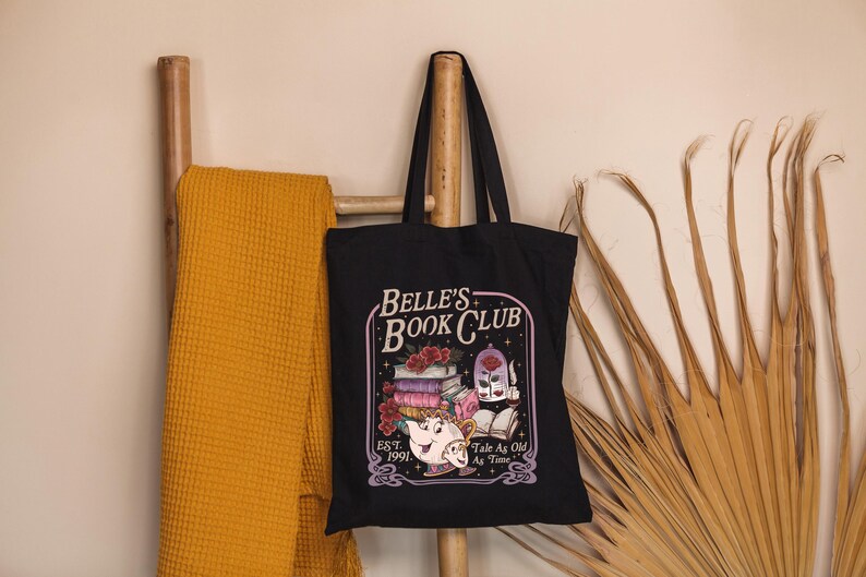 May include: Black tote bag with the text "Belle's Book Club" and a graphic of books, a teapot, and a rose. The bag is hanging on a wooden ladder with a yellow blanket. Dried palm leaves are on the right.